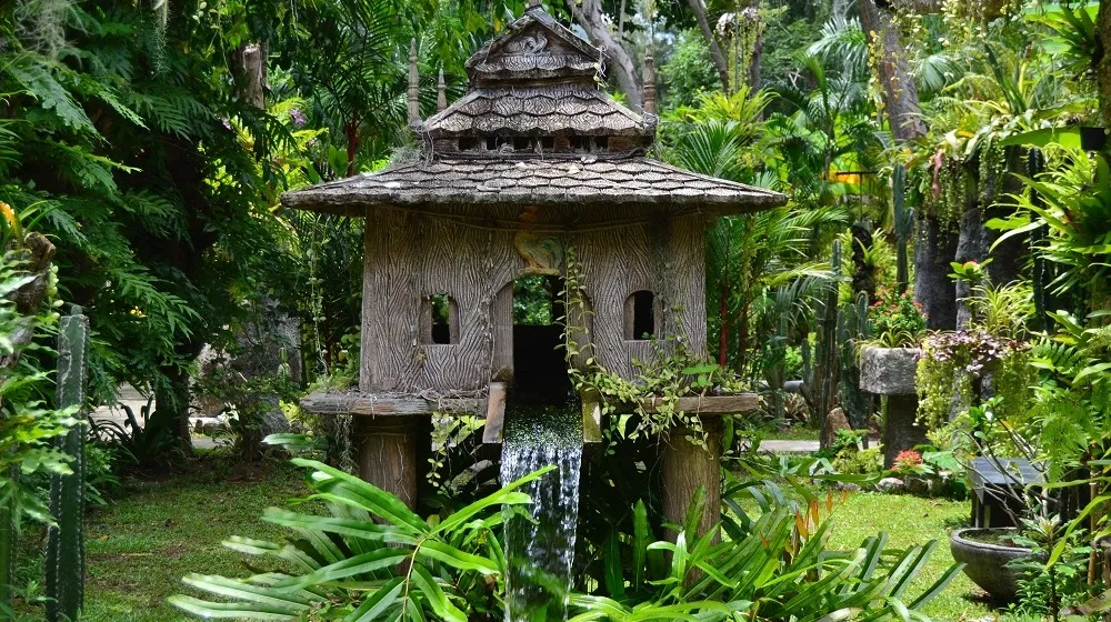 An ornate wooden spirit house beside a small waterfall in a lush tropical garden